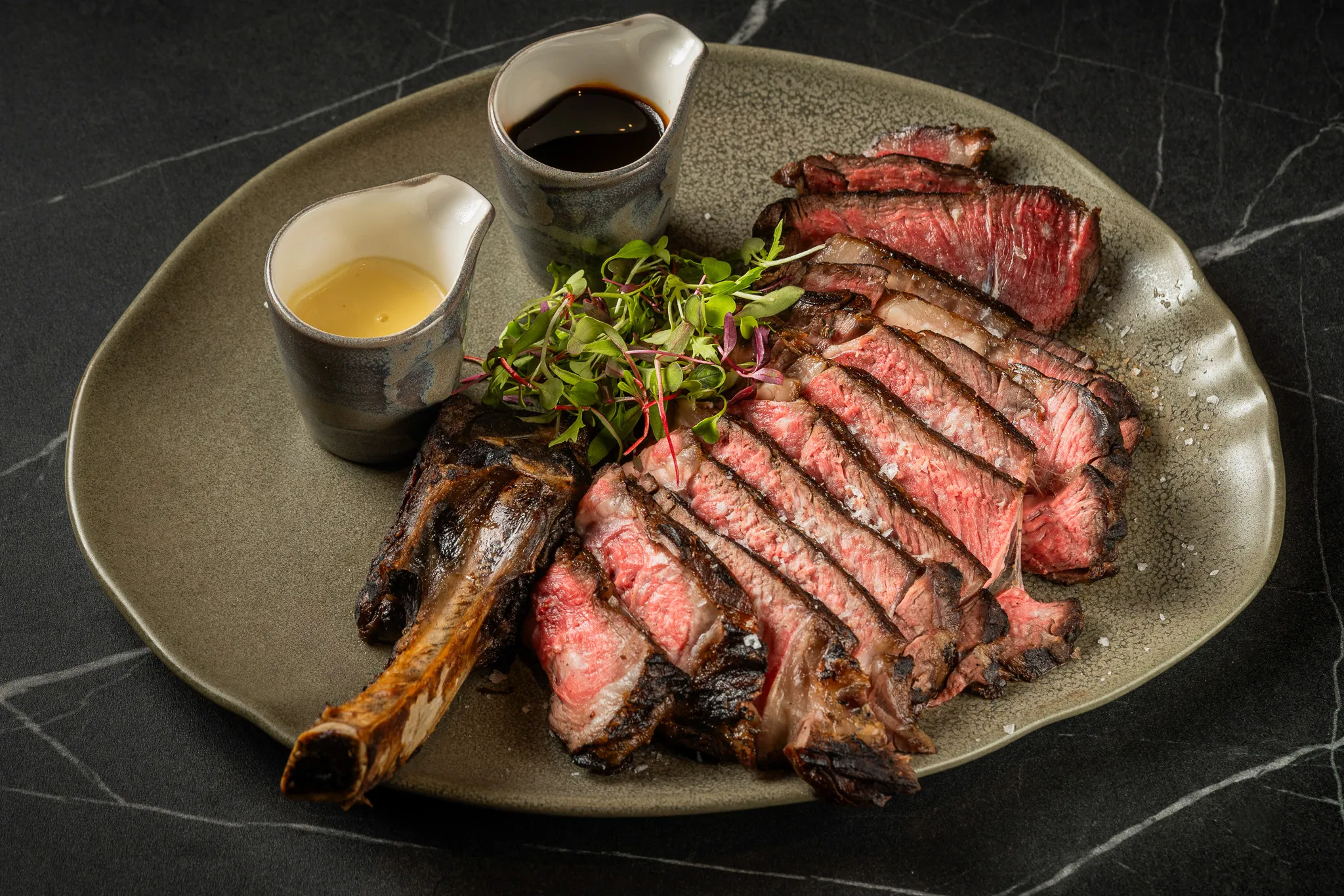 Sliced bone-in steak with greens and two sauce jugs on a ceramic platter